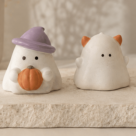 Two small white ghost-shaped decorative figurines arranged on a stone tray in a softly styled indoor setting. One ghost wears a lavender hat and holds an orange pumpkin, while the others have small orange ear or nose accents.