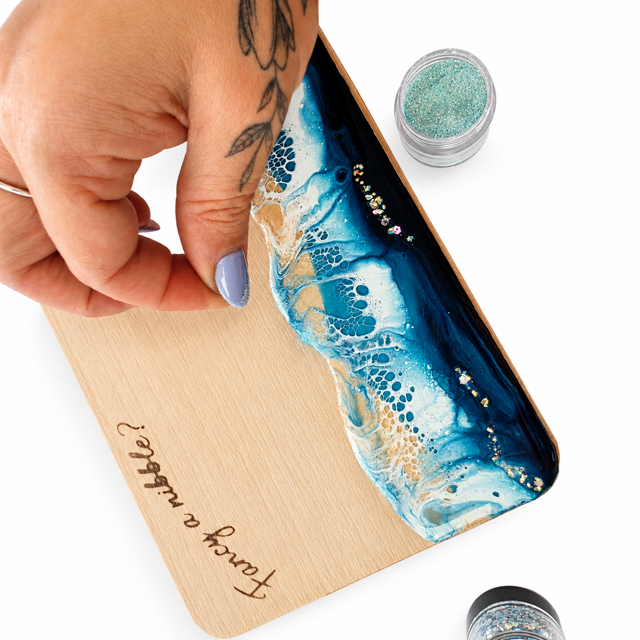 A hand with lavender nail polish is placing a rhinestone onto a rectangular wooden board with a blue, white, and gold fluid acrylic resin design. The board is engraved with the words 'Fancy a nibble?' and is shown on a clean white background with glitter jars nearby.