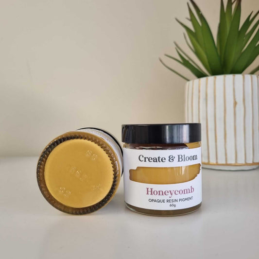 A jar of Opaque Resin Pigment labeled 'Honeycomb' with a pastel orange color swatch on the lid, placed next to a white vase with a green plant.
