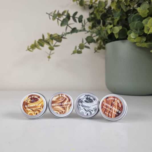 Four small containers of metallic mica pigment powder arranged in a row with a plant in the background, used for creating metallic effects in resin art and crafts.