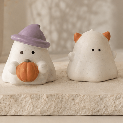Two small white ghost-shaped decorative figurines arranged on a stone tray in a softly styled indoor setting. One ghost wears a lavender hat and holds an orange pumpkin, while the others have small orange ear or nose accents.