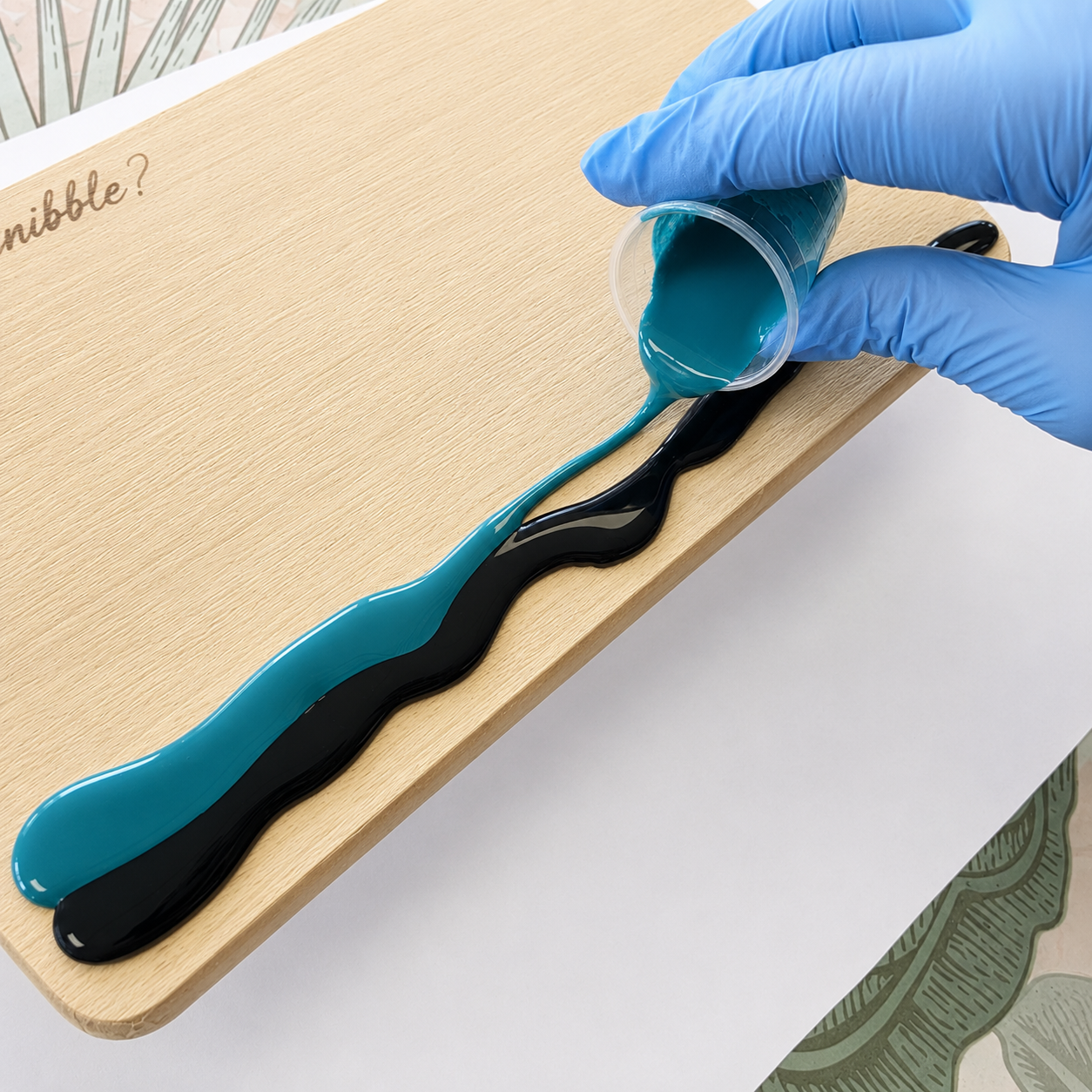 A hand wearing a blue nitrile glove pours teal and black liquid resin in a wavy pattern onto a light wood board. The resin flows from a small clear plastic cup.