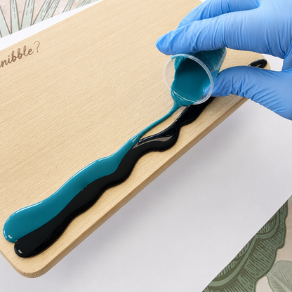 A hand wearing a blue nitrile glove pours teal and black liquid resin in a wavy pattern onto a light wood board. The resin flows from a small clear plastic cup.
