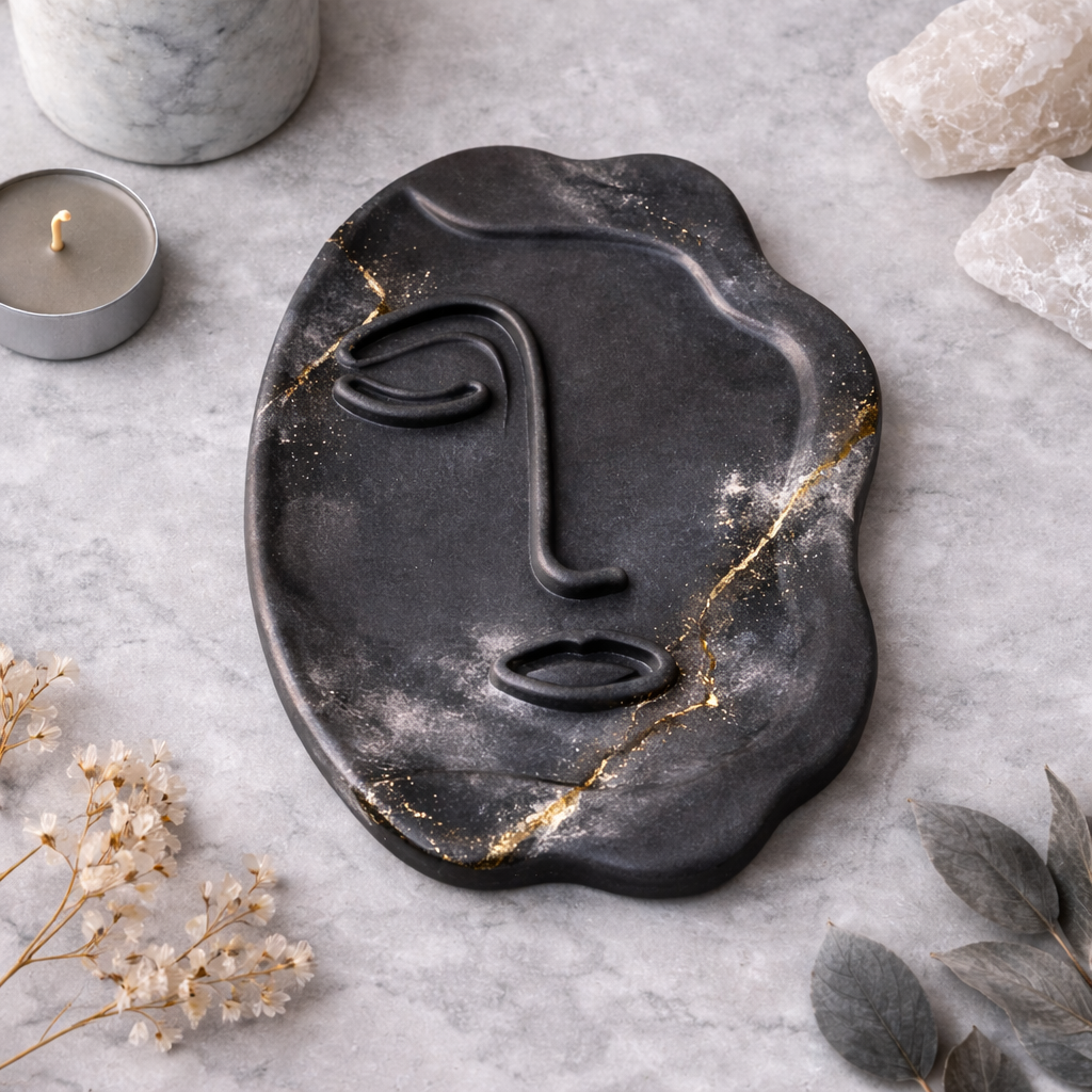 Black marble face-shaped tray with gold accents on a gray surface with candles and crystals.