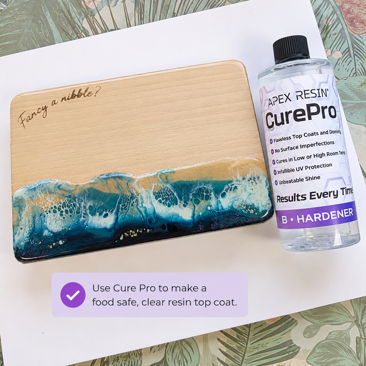 A wooden board with a blue and teal resin wave design and the engraved phrase 'Fancy a nibble?' is shown next to a bottle of Apex Resin CurePro hardener. The setup is on a white sheet of paper with a decorative tropical leaf pattern background, and a purple text bubble explains using CurePro for a clear resin top coat.