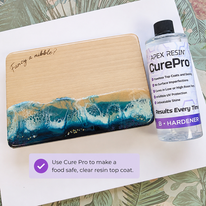A wooden board with a blue and teal resin wave design and the engraved phrase 'Fancy a nibble?' is shown next to a bottle of Apex Resin CurePro hardener. The setup is on a white sheet of paper with a decorative tropical leaf pattern background, and a purple text bubble explains using CurePro for a clear resin top coat.