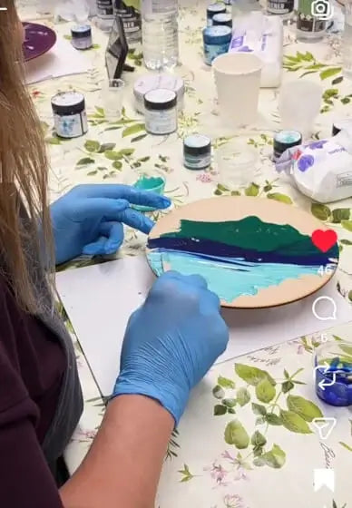Person wearing blue gloves painting a ceramic item on a table with art supplies.