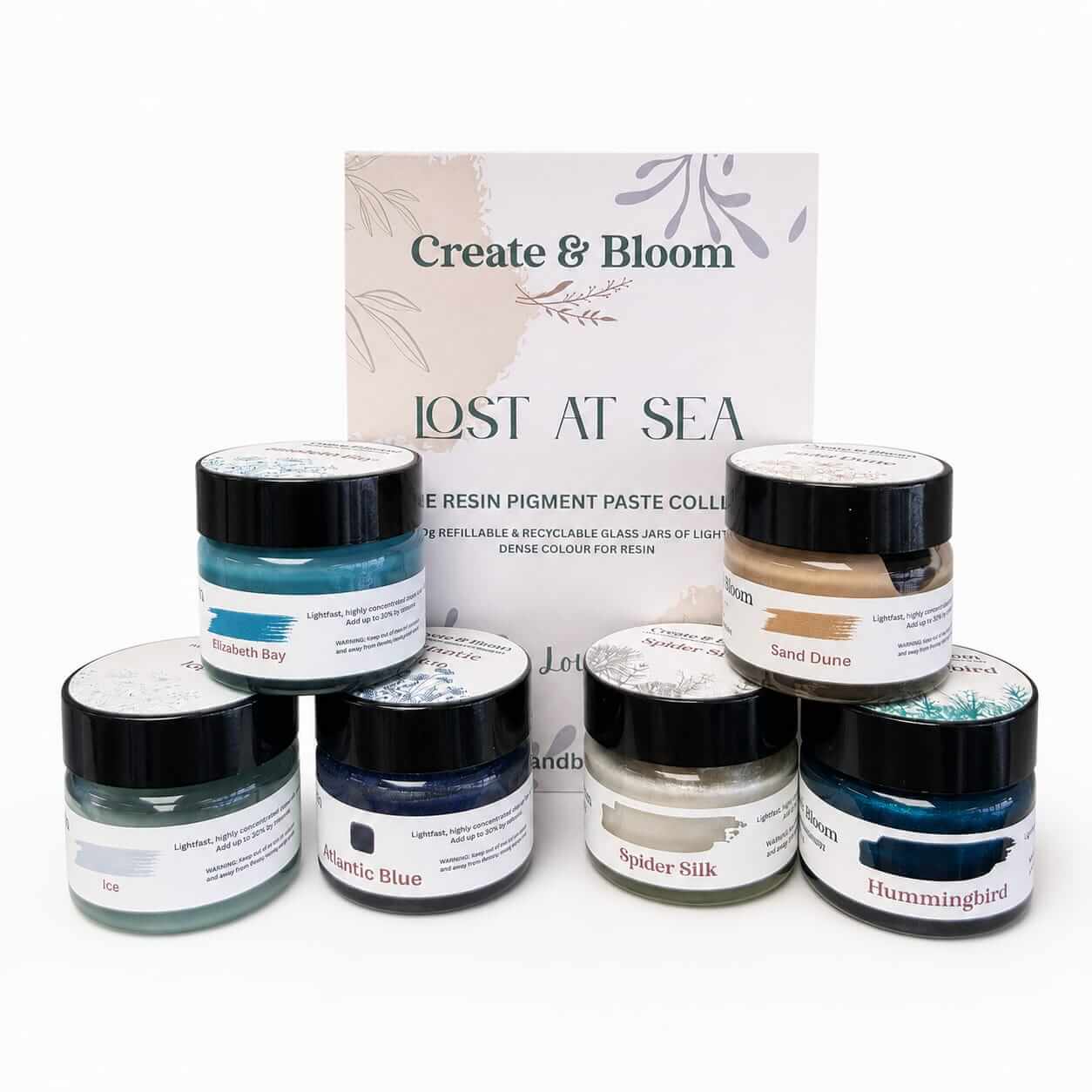 A resin pigment paste collection set by Create & Bloom, titled 'Lost at Sea', featuring six small glass jars with black lids and various paint-like pastes in blue, beige, gray, and white, arranged in front of a branded backing card on a white background.