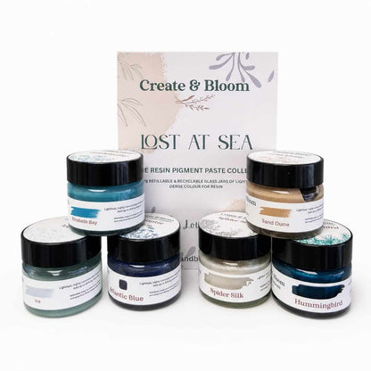 A resin pigment paste collection set by Create & Bloom, titled 'Lost at Sea', featuring six small glass jars with black lids and various paint-like pastes in blue, beige, gray, and white, arranged in front of a branded backing card on a white background.