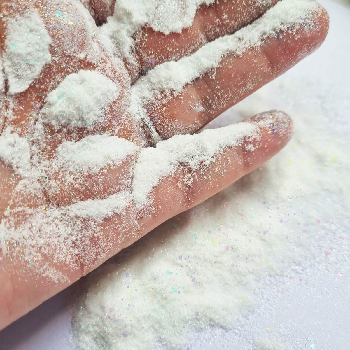 A close-up of a hand covered in fine white glittery powder, with more of the same white iridescent powder spread on a white surface below.